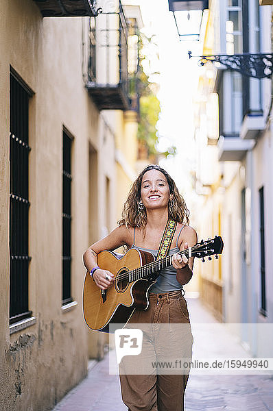 Portrait of smiling young woman playing guitar while walking on narrow street at Santa Cruz  Seville  Spain