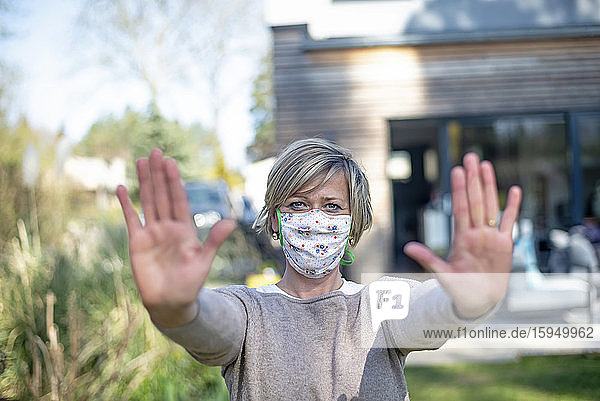 Porträt einer reifen Frau  die eine schützende Gesichtsmaske trägt  während sie während der Abriegelung der Epidemie mit einer Stopp-Geste gegen das Haus steht