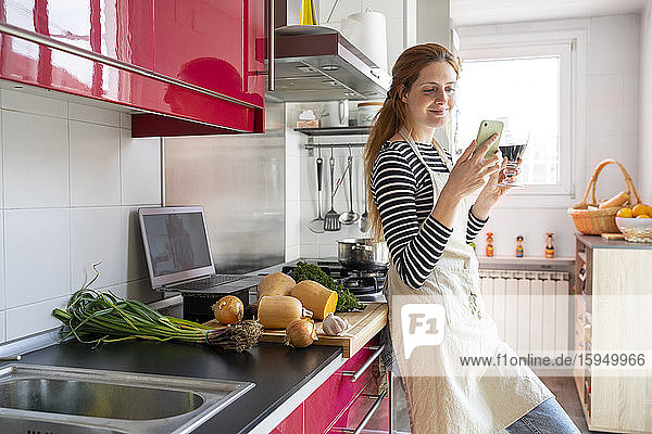 Frau trinkt ein Glas Rotwein in der Küche,  während sie ein Smartphone benutzt