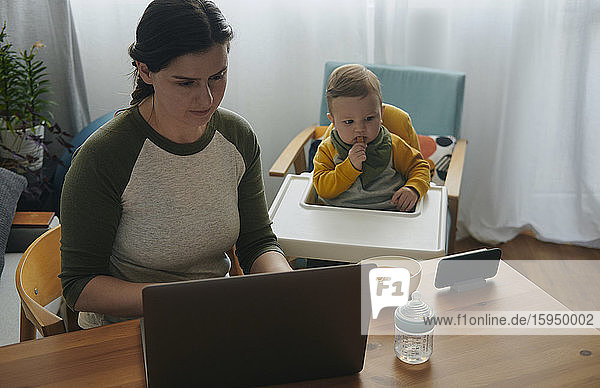 Frau arbeitet zu Hause am Laptop  während ihr kleiner Sohn Filme auf dem Handy anschaut