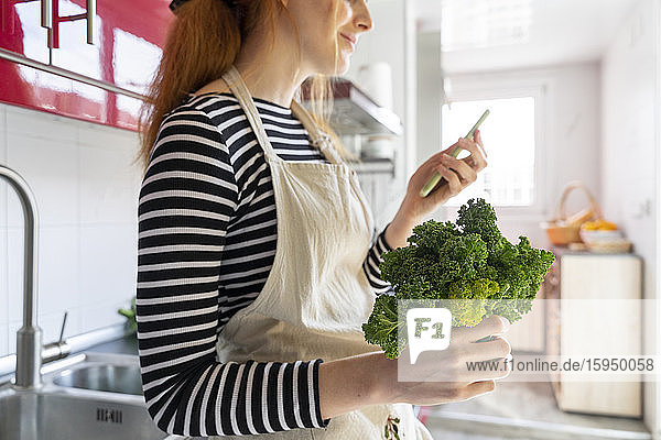 Junge Frau steht in der Küche  hält Grünkohl und benutzt ein Smartphone