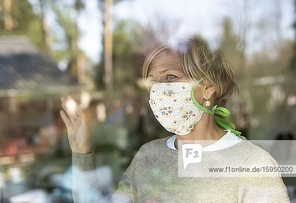 Reife Frau mit Gesichtsmaske  die während der COVID-19-Pandemie durch Fensterglas schaut