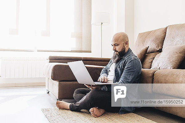 Businessman with shaved head working on laptop while sitting on floor against sofa at home