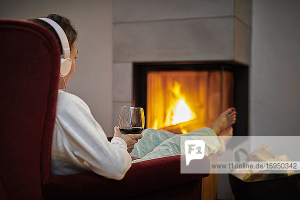 Frau sitzt auf einem Sessel mit einem Glas Rotwein und hört Musik mit Kopfhörern vor dem Kamin