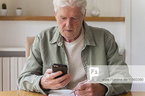 Pensionierter zuckerkranker älterer Mann benutzt Smartphone  während er zu Hause mit seinem Tagebuch sitzt