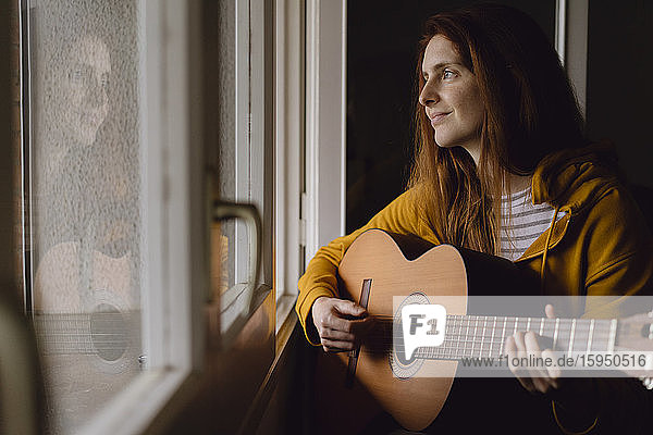 Porträt einer entspannten rothaarigen Gitarre spielenden Frau am offenen Fenster