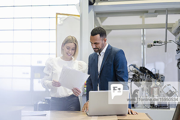 Businessman and young woman with papers and laptop talking in a factory