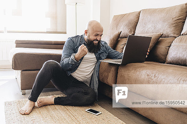 Happy businessman cheering with clenching fist while looking at laptop in living room