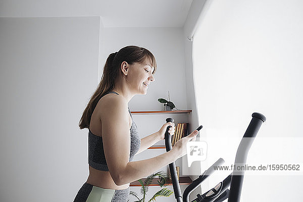 Frau benutzt ein Smartphone  während sie zu Hause auf einem Ellipsentrainer trainiert