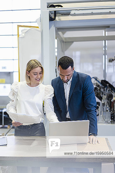 Geschäftsmann und junge Frau mit Papieren und Laptop unterhalten sich in einer Fabrik