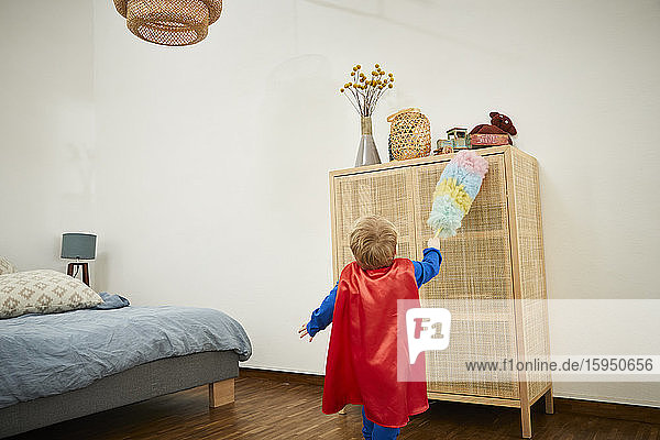 Junge in Superman-Kostüm  der zu Hause auf einer Anrichte mit Staubwedel putzt