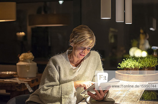 Lächelnde blonde Frau arbeitet spät  während sie ein digitales Tablet im beleuchteten Wohnzimmer benutzt