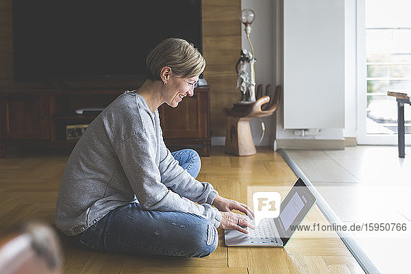Reife Frau  die von zu Hause aus arbeitet  einen Laptop benutzt und im Schneidersitz auf dem Boden sitzt