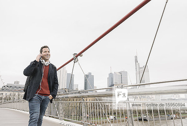 Lächelnder Geschäftsmann telefoniert mit einem Mobiltelefon  während er auf einer Brücke gegen den Himmel in Frankfurt geht