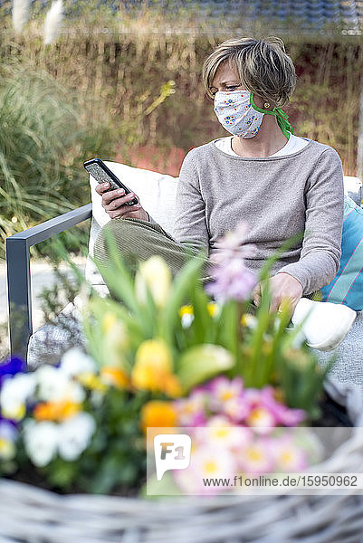 Frau trägt Gesichtsmaske und benutzt ein Smartphone  während sie während der Abriegelung auf der Terrasse sitzt