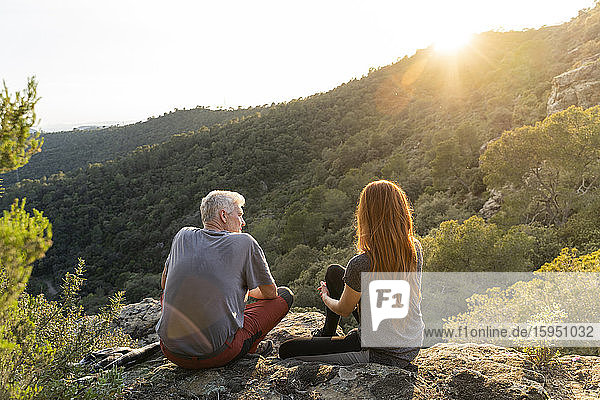 Rückenansicht eines Vaters und einer erwachsenen Tochter  die auf einem Felsen in den Bergen sitzen und den Sonnenuntergang genießen