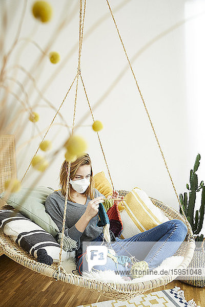 Blonde Frau trägt FFP2-Maske und strickt zu Hause im Hängestuhl sitzend