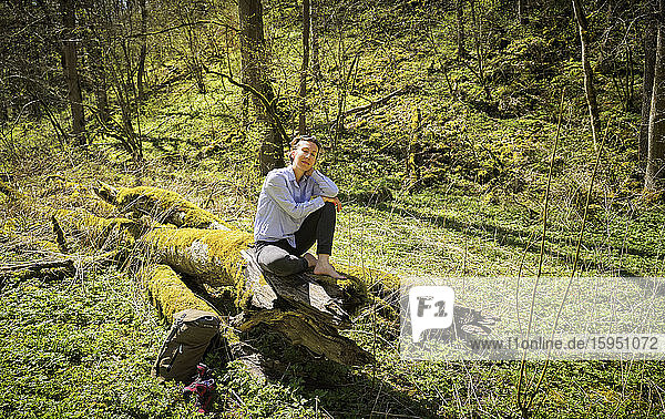 Entspannte Frau in voller Länge auf moosbedecktem Baumstamm im Wald der Schwäbischen Alb sitzend