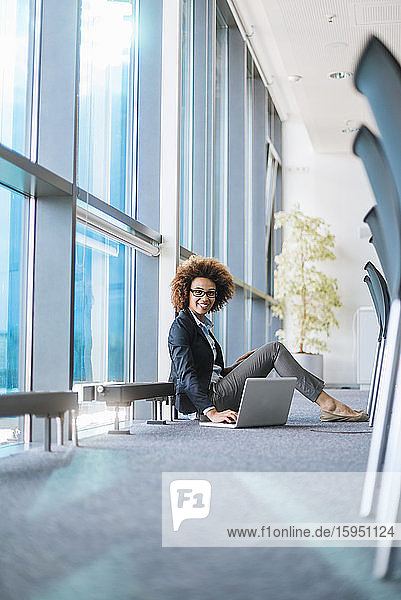 Lächelnde junge Geschäftsfrau sitzt mit Laptop auf dem Boden im Konferenzraum