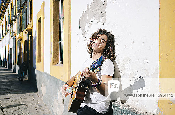Lächelnder junger Mann mit geschlossenen Augen spielt Gitarre  während er an einer Wand lehnt  in Santa Cruz  Sevilla  Spanien