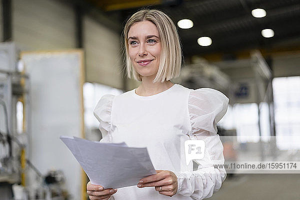 Porträt einer lächelnden jungen Frau  die in einer Fabrik Papiere in der Hand hält
