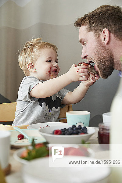 Porträt eines verschmierten kleinen Jungen  der seinen Vater am Frühstückstisch mit Marmeladenbrot füttert