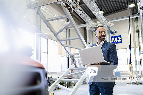 Smiling businessman holding laptop in a factory