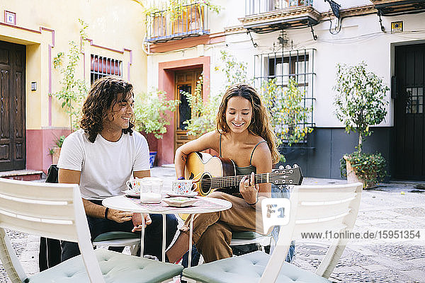 Lächelnder junger Mann sieht seine Gitarre spielende Freundin an  während er in einem Straßencafé sitzt  Santa Cruz  Sevilla  Spanien