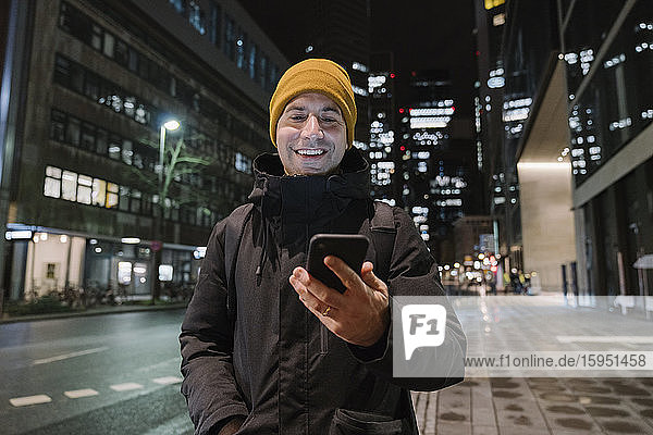 Porträt eines lächelnden Mannes  der nachts auf ein Smartphone schaut  Frankfurt  Deutschland