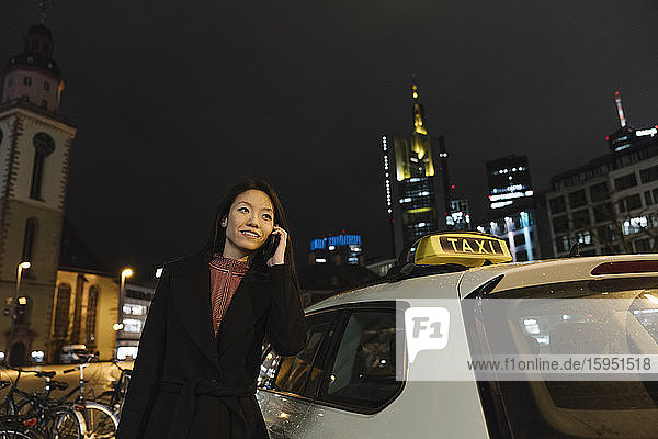 Junge Frau am Telefon neben einem Taxi in der Stadt bei Nacht  Frankfurt  Deutschland