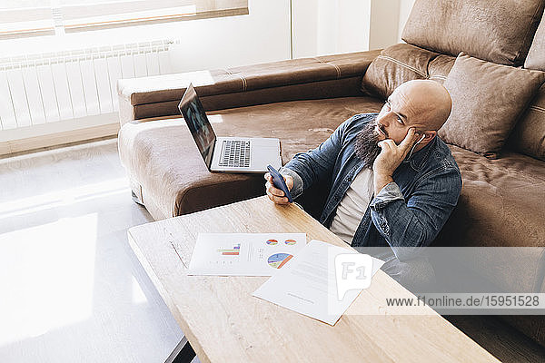 Nachdenklicher Geschäftsmann mit Laptop und Dokumenten mit Smartphone im Wohnzimmer