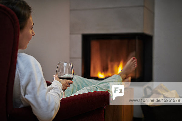 Woman sitting on armchair with glass of red wine relaxing in front of fireplace