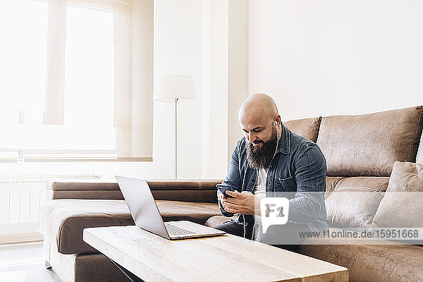 Mann hört Musik über Mobiltelefon  während er zu Hause am Laptop arbeitet