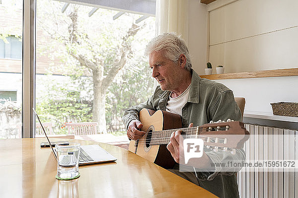 Retired senior man learning to play guitar through online tutorials on laptop at home