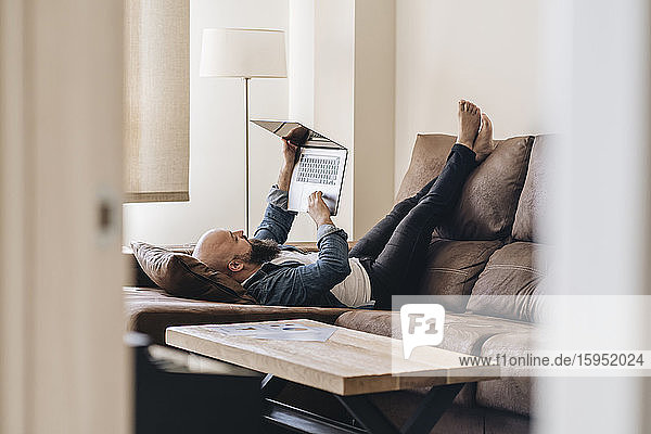 Gelangweilter Mann benutzt Laptop  während er zu Hause auf dem Sofa im Wohnzimmer liegt