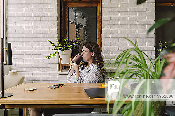 Nachdenkliche Frau trinkt Kaffee  während sie im Heimbüro am Schreibtisch sitzt