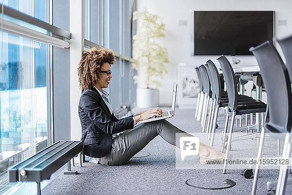 Smiling young businesswoman sitting on the floor in conference room using laptop