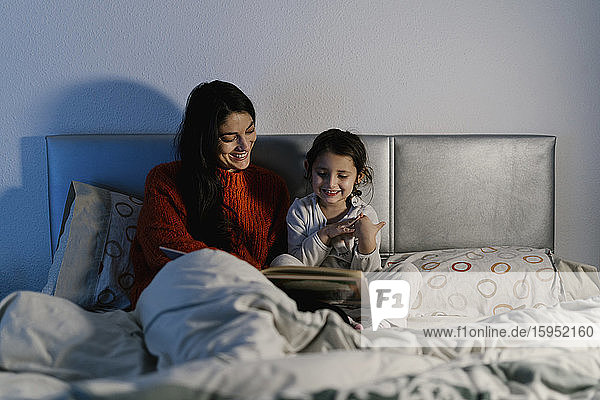 Glückliche Mutter und ihre kleine Tochter sitzen zusammen im Bett und schauen Bilderbuch