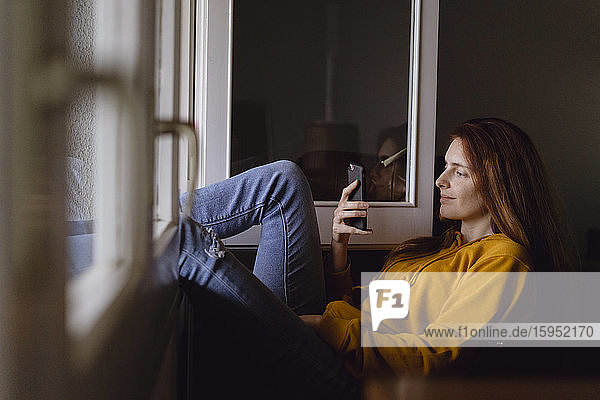 Lächelnde rothaarige Frau sitzt mit erhobenen Füßen am offenen Fenster und schaut auf ihr Handy