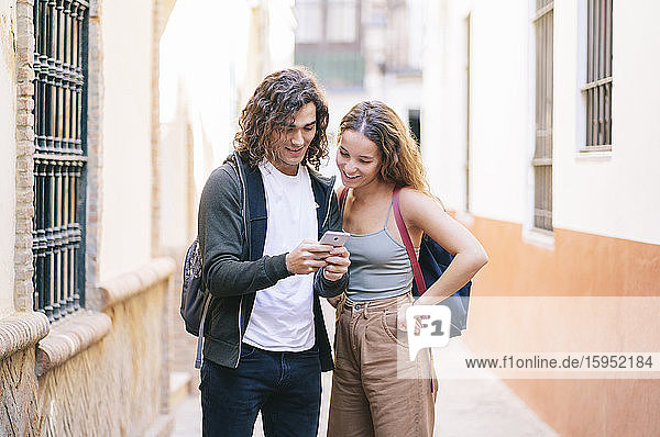 Lächelnde junge Frau  die einen Mann mit einem Smartphone ansieht  während sie in einer engen Straße in Santa Cruz  Sevilla  Spanien  steht