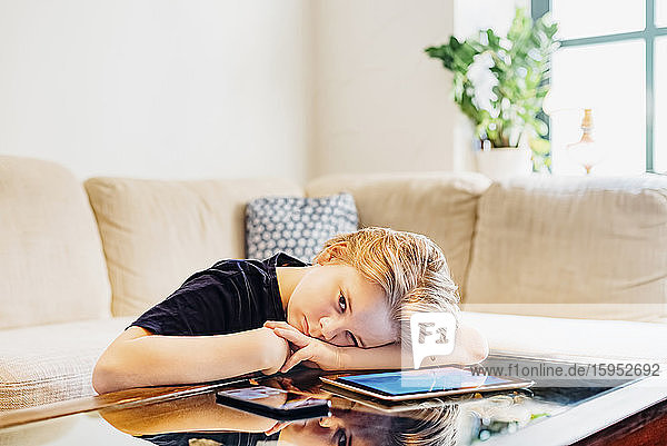 Serious boy lying on coffee table with smartphone and tablet