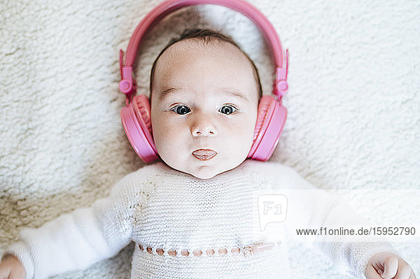 Porträt eines kleinen Mädchens mit übergrossen rosa Kopfhörern  die auf einer Decke liegen und die Zunge herausstrecken