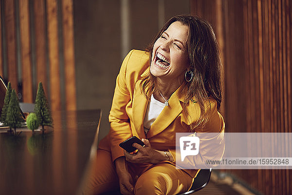 Lachende Geschäftsfrau im gelben Anzug sitzt am Schreibtisch im Büro