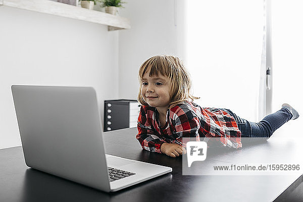 Little girl lying on desk looking at laptop