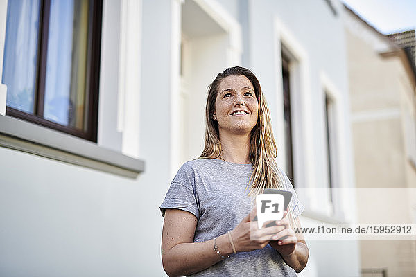 Woman looking away while holding mobile phone against house