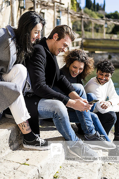 Vier glückliche Freunde sitzen auf einer Treppe am Gardasee und teilen sich ein Smartphone  Italien