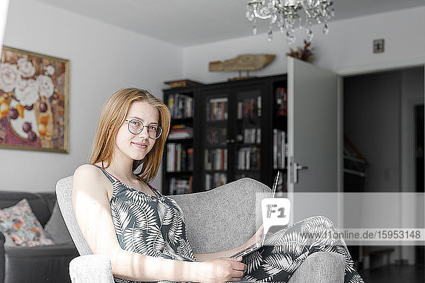 Porträt einer lächelnden jungen Frau mit Laptop  die auf einem Sessel im Wohnzimmer sitzt