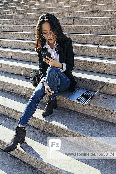 Junge Geschäftsfrau mit Laptop sitzt auf einer Treppe im Freien und schaut auf ihr Handy
