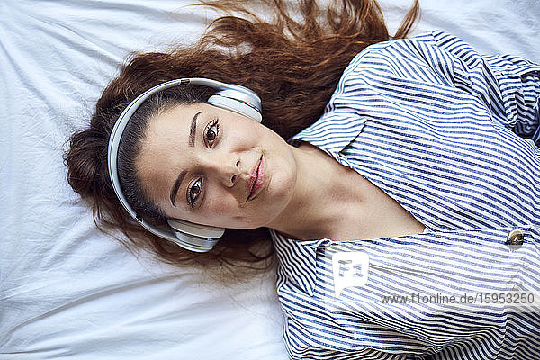 Porträt einer lächelnden jungen Frau  die auf dem Bett liegt und mit Kopfhörern Musik hört