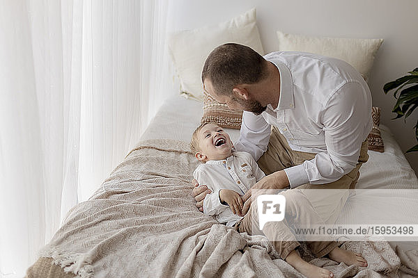 Vater kauert auf dem Bett und kitzelt seinen kleinen Sohn